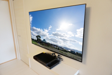 「白」の多い明るい雰囲気のリビングにブラビア・55インチ「KJ-55X8500E」の壁掛けとブルーレイレコーダー用の専用シェルフを同時に施工させて頂きました。

オーナー様は壁掛けテレビへのキッカケに、「省スペースさ」を重視されたと仰っていました。
生活動線上で、テレビ台を設置してしまうとどうしても狭さを感じてしまう点と、掃除のしやすさも意識されてご依頼されたとお伺い致しました。

事前のコンサルでは、テレビ台をどうしても置きたくないので、周辺機器の置き場所に悩まれていましたが、専用のシェルフをご提案させて頂く事で、解決致しました。

リビングへの入口から正面にテレビの側面が見えてしまう事もあり、テレビに合わせて金具を薄型タイプに選定し、テレビの壁からの出幅も大きく抑えて仕上げさせて頂きました。

シェルフ出幅も25cmとテレビ台と比較してもスッキリとした仕上がりに大きく貢献できています。

施工後には求められていた省スペースさを強く実感して頂く事が出来まして、見た目のカッコよさにもご夫婦から喜びの声も頂くことができ、大変嬉しく思います。