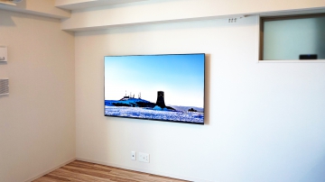 リビングの広さを最大限に活かして、ソニー・ブラビア有機ELテレビ「KJ-65A9G」を壁掛け施工させて頂きました。

オーナー様は新築マンションへの引っ越しをきっかけに、テレビの買い替えと壁掛けテレビをイメージされて、テレビの購入から壁掛け施工までを当店で一貫してご用命頂く事ができました。

リビングの印象をよりスッキリとスタイリッシュに見せたいという想いから、壁からの出幅を最大限に抑えられるテレビと金具の組み合わせをご提案させていただきました。

ポータブルタイプの外付けHDDをテレビ裏に隠して設置することで、場所を取る事無く録画機能も楽しむ事ができ、ゲーム機やブルーレイディスク・DVDの再生の事を考慮して遊びたいときにだけ接続できるように、テレビ下にはHDMIコンセントも新設させて頂きました。

プレーヤー等を使いたいときにだけ接続できるようにしておく事で、使いやすさも活かしつつ、テレビだけが壁掛けされている理想的な空間にも出来る事が今回の組み合わせの最大の魅力と考えます。

施工後には、有機ELテレビの美しい映像とテレビだけのスタイリッシュな空間にとても喜んで頂く事が出来ました。